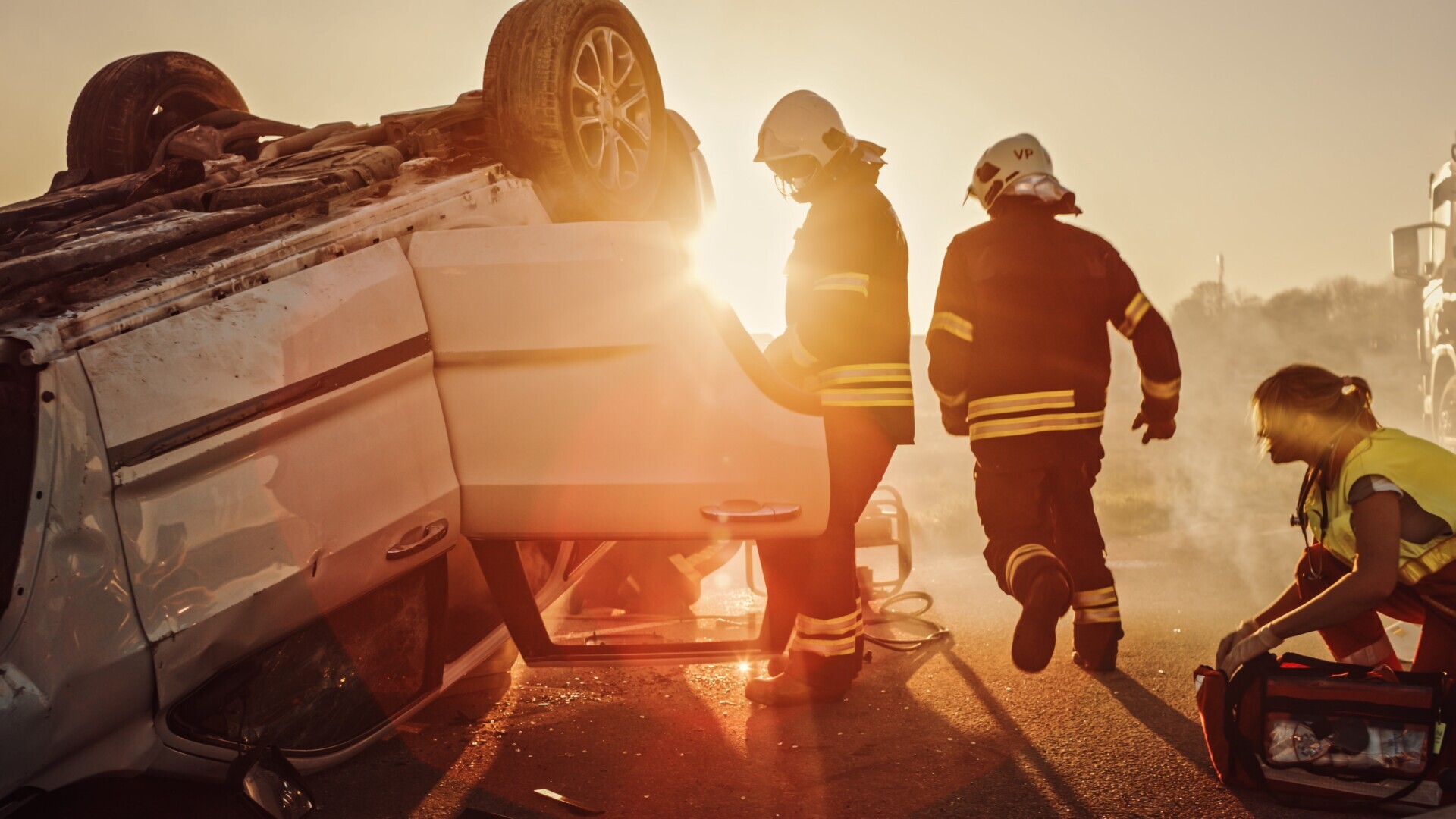 Firefighters assess an overturned vehicle at sunrise, while an emergency responder examines equipment nearby.