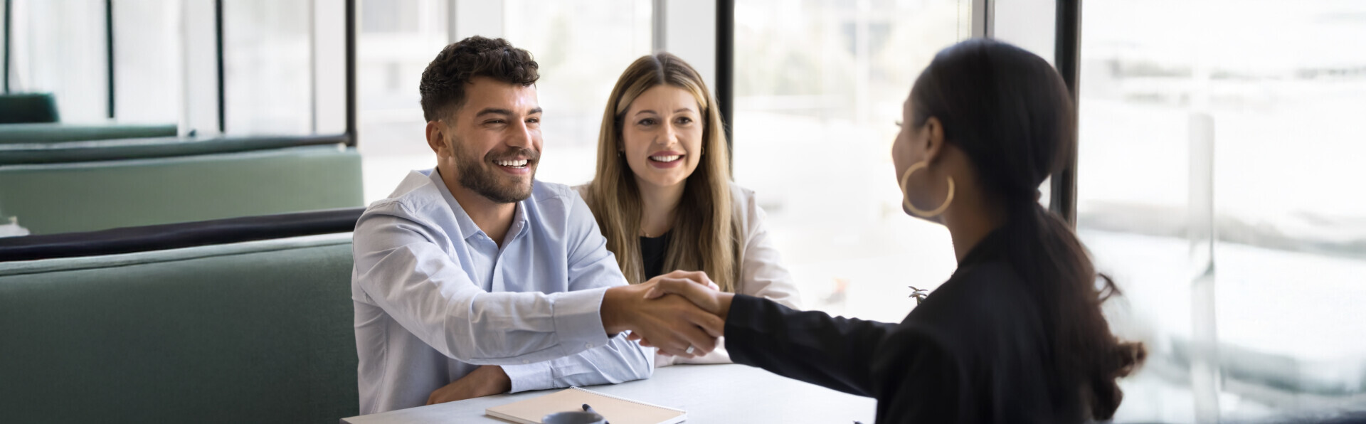Three people at a table in a well-lit office space. Two people are seated, smiling, and shaking hands with a third person who is standing.