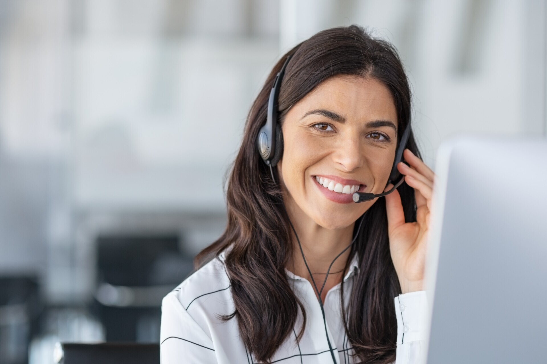 A woman with long brown hair wearing a headset smiles while sitting at a desk in an office. A computer monitor is partially visible in front of her.